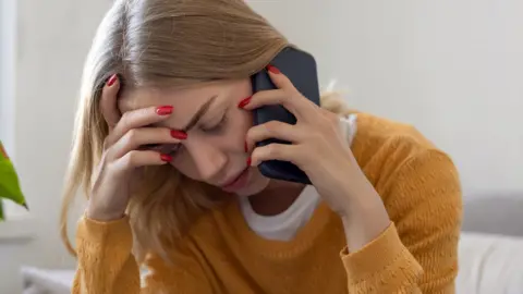 Woman wearing a yellow sweater has her hands on her head while holding up a phone to her ear. She looks stressed.