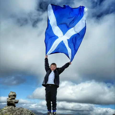 HANNAH ANDERSON A boy with dark hair standing on top of a mountain. He is holding a Saltire above his head which is fluttering in the wind against a grey sky. There is a small pile of stones in the background.