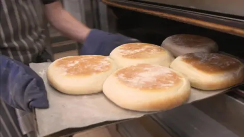 A baker takes five freshly-baked stotties out of the oven.