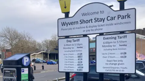 A car park sign reads "Welcome to Wyvern short stay car park" and includes information on parking tariffs. Parked cars and trees can be seen in the background and there is a black payment machine to the left of the sign. 