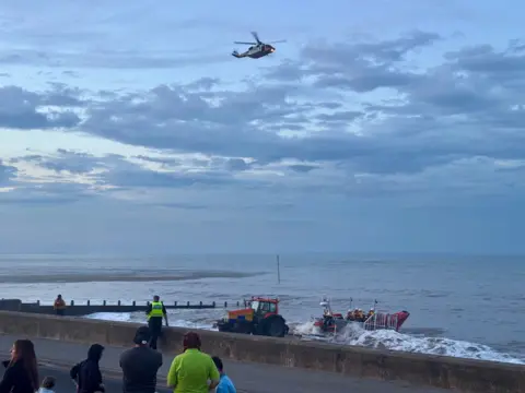 Phillip Norton/BBC An orange search and rescue boat is launched into a the sea from a beach. An expanse of water, helicopter and grey sky are visible overhead.