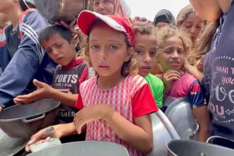 Young children wait in a line for food, looking anxious, holding empty pots in a crowded scene.