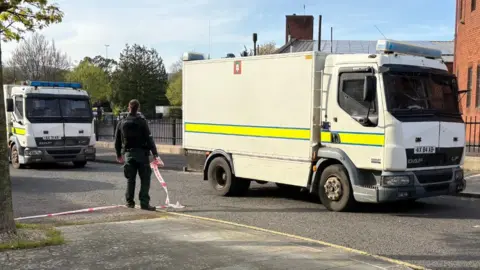 Two large white police trucks parked in front of a red brick building. A police man, dressed all in black, is stood between the trucks, back to camera, holding white and red cordon tape in a residential street. There are trees and railings visible in the background.