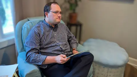 Nicholas Strahl Nicholas Strahl, wearing glasses and a checked shirt, sits in an armchair holding a tablet.