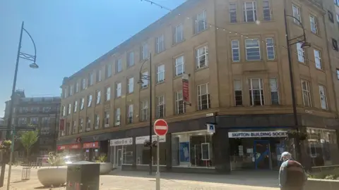 A large city centre building with four floors. A ground floor unit bears the sign 'Skipton Building Society'. It is a sunny day and other street furniture can be seen.