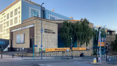 A sandstone building with black lettering spelling the University of Bradford with a pedestrian crossing in front. 