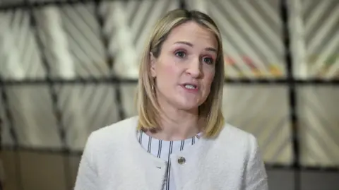 Helen McEntee is talking. She has shoulder-length blonde hair and is wearing a cream coloured jacket over a stripy top. Behind her out of focus a glass wall can be seen.