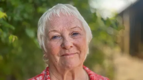 Stephen Huntley/BBC A woman with short grey hair smiles at the camera. She wears a red shirt with a white pattern on it. She is standing outside on a sunny day. The trees behind her are blurred.