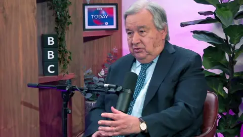 Antonio Guterres gestures as he talks into a microphone with a plant in background during an interview with BBC