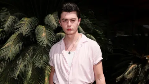 Getty Images Reay poses at a California party in front of a palm tree plant. He has a dark quiff and wears a short sleeved pink shift over a white top and chain