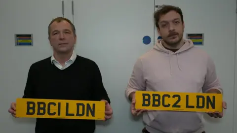 Jim Wheble and a colleague stand in an office holding customised number plates. The plates read: BBCI LDN and BBC2 LDN.