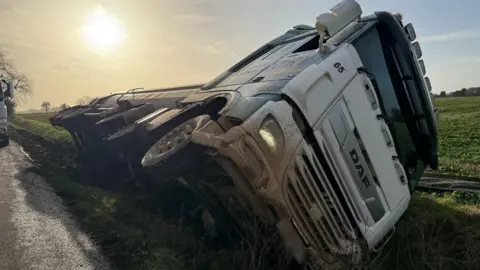 An overturned lorry is on its side in a ditch at the side of a road. The sun is shining in the sky and there are fields in the background.