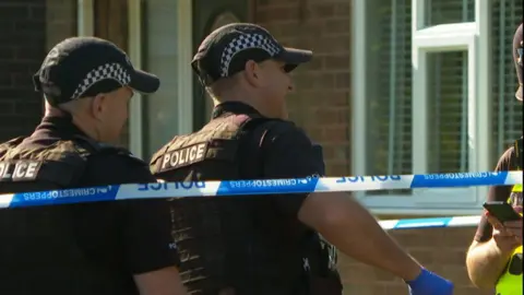 Two officers in uniform are seen working on the case behind police tape. 