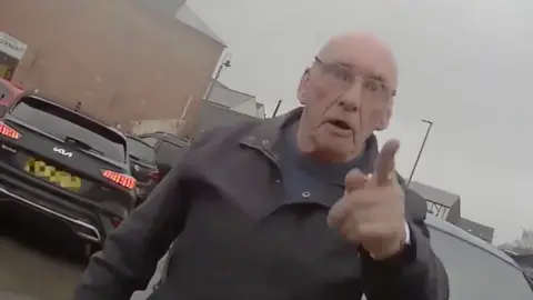 A man standing in a car park. He has his finger pointed at the person wearing the body-cam.