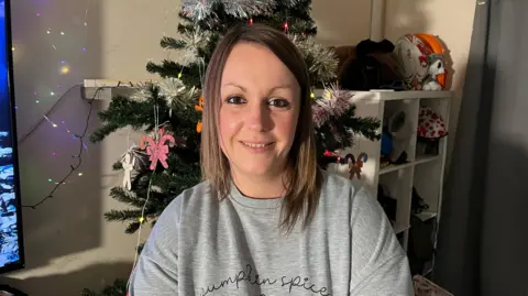 BBC A woman sitting in front of a Christmas tree and a white cabinet with toys on top and in the sections. White wall with a TV to the left.