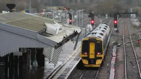 Shuan Croucher A train pulls into a railway station platform where the roof is damaged. Corrugated iron is hanging down towards the platform. 