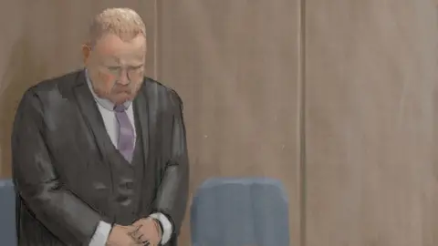 Nick Lewis Robert Bush stands in the dock at Hull Crown Court wearing a formal dark suit, white shirt and mauve tie. He has his head down and his hands clasped in front of him. 