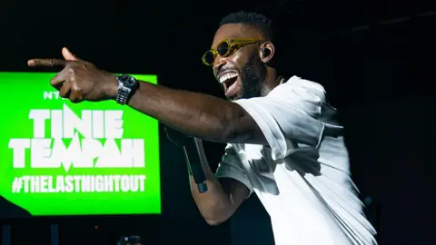 Getty Images Tinie Tempah pointing left of the camera. He's smiling and wearing sunglasses and a white t-shirt while singing into a mic. Behind him is a green screen that says "Tinie Tempah - hashtag the last night out".