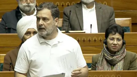SansadTV Rahul Gandhi, wearing a white t-shirt, standing in parliament with a paper in his hand. 