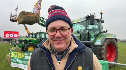 Richard Knights/BBC Tim Bradshaw wearing a read and blue bobble hat, patterned neckerchief, a beige zipped jumper and a country-style zipped gillet. He is standing in front of a tractor which has lifted a hay bale.