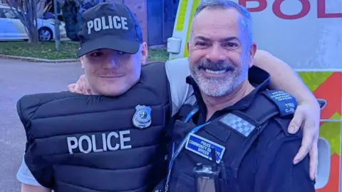 Nottinghamshire Police Ryan Sissons wearing a police cap and vest with PCSO Chris Jones in his uniform standing in front of a police van