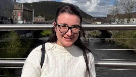Rachel Hansen stands in front of the river Taf in Pontypridd wearing a white jumper. She had dark hair and black-rimmed glasses. It is a head and shoulders shot.