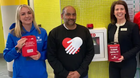 BBC (L-R) Laura Hughes, Nikhil Misra and Suzanne Grant pose at a Merseyrail station holding bleed control kits