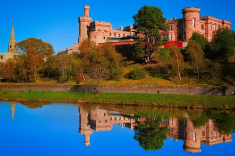 Getty Images Inverness Castle is a red sandstone building with mock turrets and battlements. The castle is reflected in the mirror-like water of the river below.