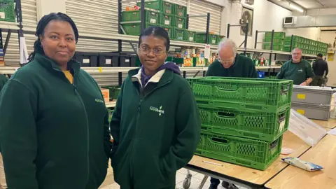 A picture of two women standing up, wearing green fleeces. In the background are three men who are packing food away into crates.