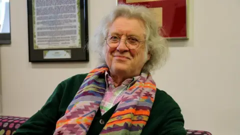 An older man wearing circular clear-framed glasses. He is sitting on a purple, spotted sofa and is wearing a purple and orange, aztec scarf. He has grey hair and is smiling into the camera. 