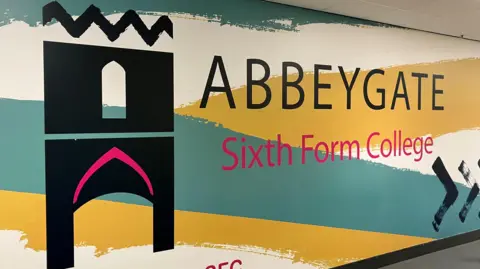 Jon Wright/BBC A large, stylised logo for the college featuring the town's medieval gateway. It is painted on a wall and includes the words "Abbeygate Sixth Form College" on a white backing with thick yellow and green stripes.