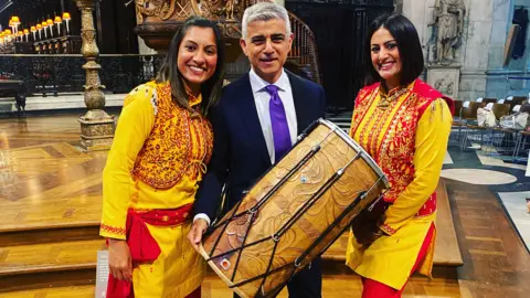 Two women with black hair stand either side of a man smiling. They are wearing yellow and red outfits with leaf and vine patterns on the tops, and red ties around their waists. They are holding a large wooden dhol drum between them. The man in the middle has grey hair and is wearing a black suit white shirt and purple tie 