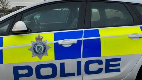 The side of a marked Lincolnshire Police car which is white with yellow and blue squares with police on the side and the force's badge