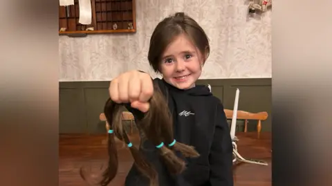 Contributed Reuben smiles at the camera while holding one arm out in front of him as he holds on to his brown hair that was cut. He wears a black hoodie as he sits on a wood table.