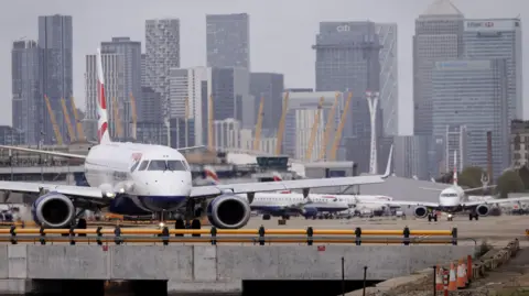 A British Airways plane sitting at the airport. The tall buildings of the Canary Wharf financial district can be seen in the background.