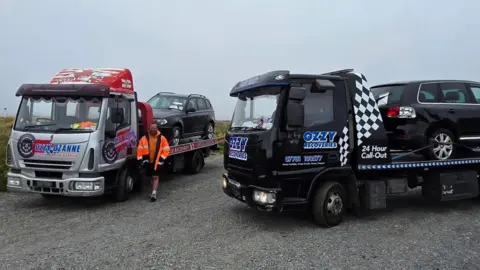 Deputy Andy Cameron Two large black SUV style vehicles on the back of two recovery trucks. The picture shows them parked in a coastal car park.