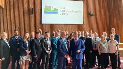 Laura Coffey/BBC Nigel Farage with short white hair in a blue suit standing next to Martin Griffiths in front of a group of councillors, many of whom are in suits. There is a white North Northamptonshire banner behind them on a wall of wooden slats.