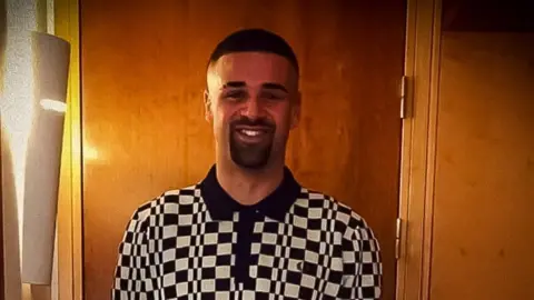 Tariq Evans, who has a shaved head and eyebrow, has a dark beard and moustache and is wearing a black and white check polo shirt