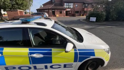 A white, yellow and blue police care dominates the front of the picture. In the background a brown brick building can be seem. There is a white sign but it cannot be read. 