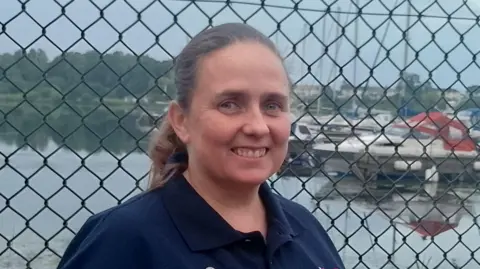 A woman with long brown hair, tied back. She is wearing a navy t-shirt. She's standing in front of a fence, with Lough Neagh visible in the background. 