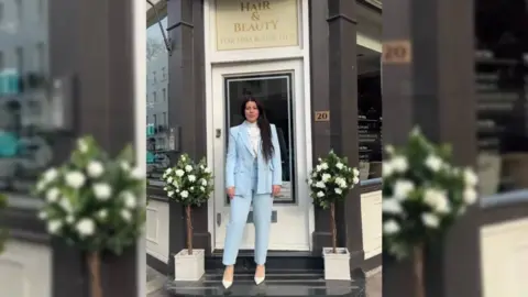 Nadia Suliaman Nadia Suliaman, has a baby blue suit on and looks directly at the camera, standing on the doorway of her salon shop in between two white rose bushes. She has dark brown hair and brown eyes. 