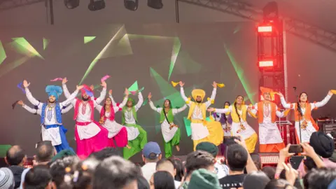 Sandwell & Birmingham Mela A group of dancers in multi-coloured outfits dance on a stage.