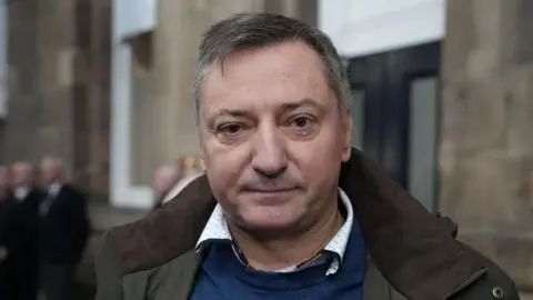 Stuart Prandle/BBC A man with short grey hair wearing a blue jumper and green and brown coat standing on a railway platform