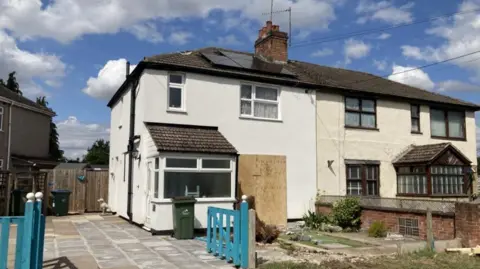 A white semi-detached house with a dark roof and a blue gate leading to a paved drive. Part of the front of the house is covered with wood.