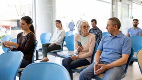 A hospital waiting room with six patients sitting in rows of blue seats.