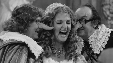 Hulton Archive via Getty Images Penelope Keith on Morecambe and Wise. They are dressed as if they were members of the French court and the two men are wearing comedy noses. Keith is laughing.