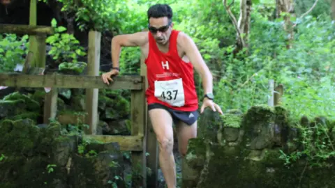 A man wearing a red running vest jumping through a stile