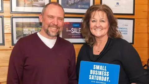 Small Business Saturday UK Karen and Dave Meadows, founders of PUSHED, stood in a wooden room holding a blue poster with the words 'small business Saturday' printed on it. Karen is brunette, and is wearing a black dress, whilst Dave is wearing a burgundy V-neck jumper and blue trousers.