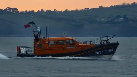 The R&J Welburn lifeboat from Exmouth RNLI. It is orange and blue. It is sailing past a section of coast during the early evening as the sun sets.