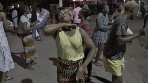 Seorang wanita dengan peluit mengangkat tangan di dagunya - di sekelilingnya terdapat pengunjuk rasa lain yang memegang pot, botol atau peluit di jalan pada malam hari di Maputo, Mozambik